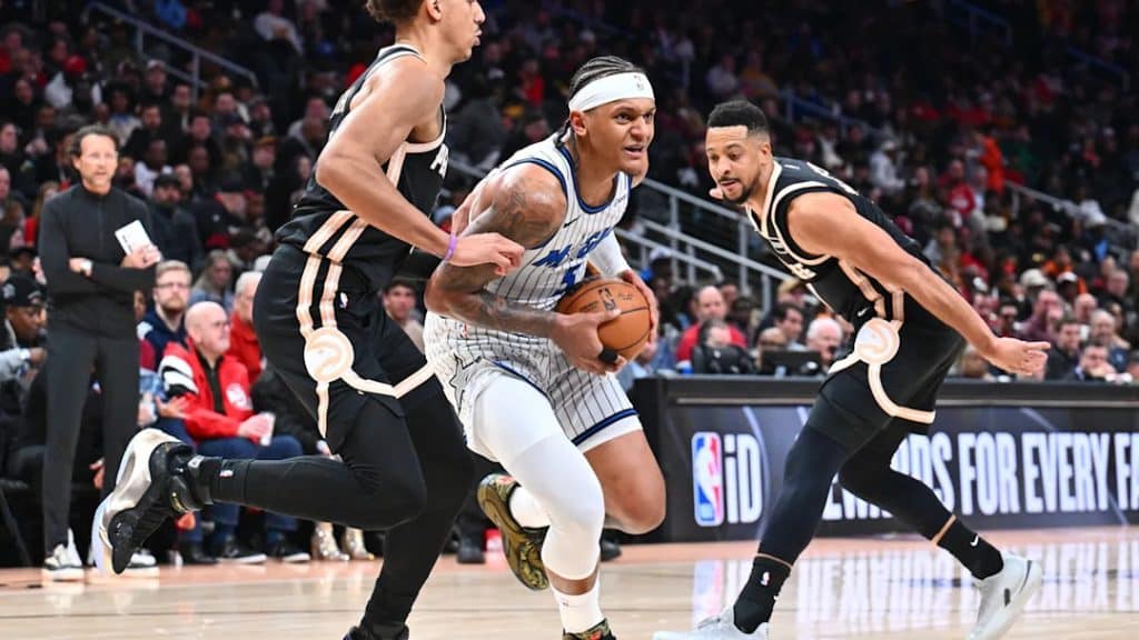 magic game 2 1 - Digital Wager Wire Basketball player in a white uniform drives with the ball while two defenders in black uniforms guard him on a crowded arena floor.