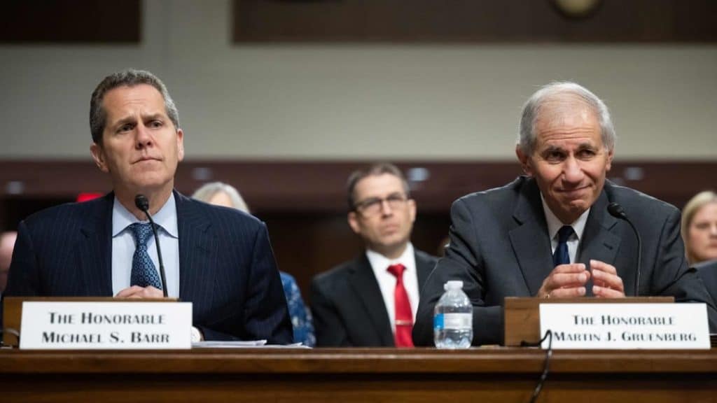 regulators 1 - Digital Wager Wire Two officials in dark suits sit at a panel table during a formal hearing, microphones in front and nameplates reading The Honorable Michael S. Barr and The Honorable Martin J. Gruenberg.