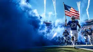 american-football-texas-longhorns-football-playersjpg - Digital Wager Wire Football player wearing #27 carries an American flag as blue smoke billows and teammates run onto the field.