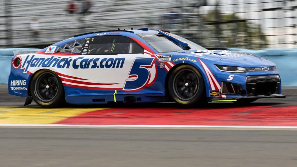 cars - Digital Wager Wire Race car in blue, white, and red livery with 'HendrickCars.com' on the side, number 5, racing on track barriers in the background