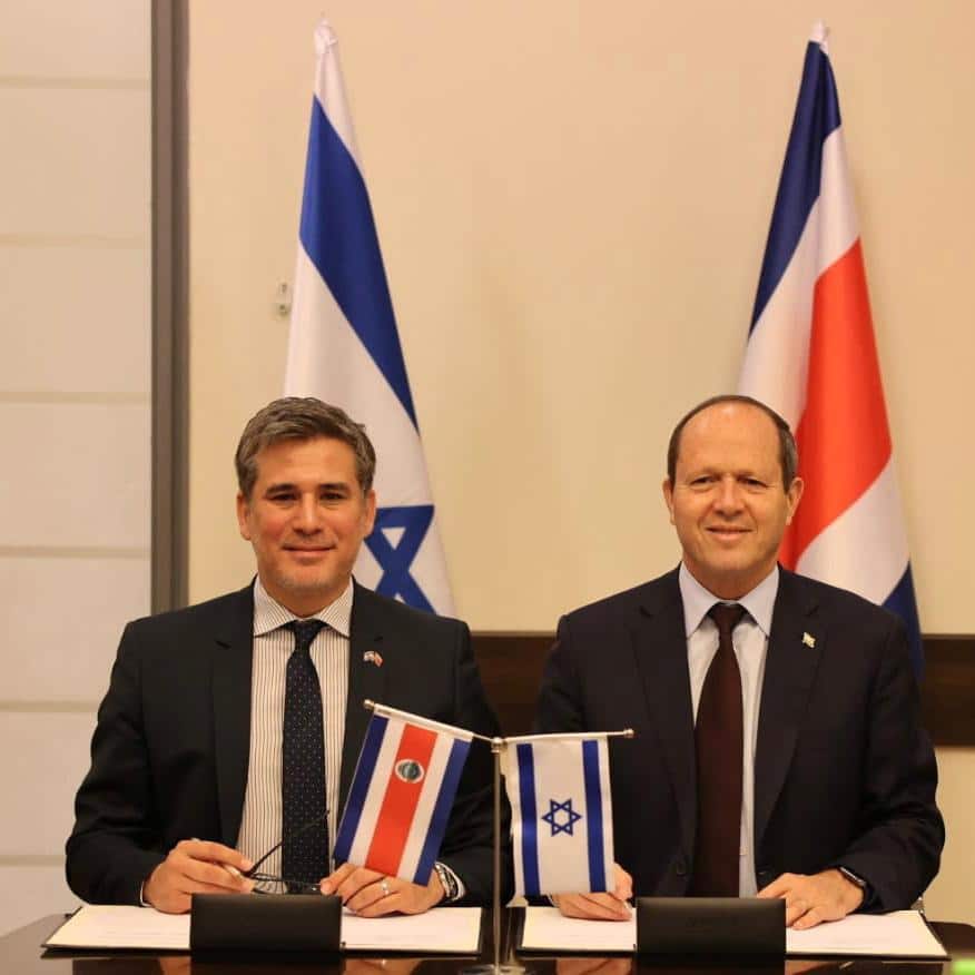 costa rica trade - Digital Wager Wire Two men in suits sign documents at a formal signing table with small Israeli and Costa Rican flags between them.