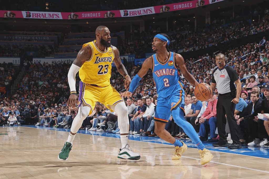 lebron defending - Digital Wager Wire Two basketball players compete for the ball on a crowded court, one in a yellow Lakers uniform (number 23) and the other in a blue Thunder uniform (number 2) as they move toward the basket with a referee and spectators in the background.
