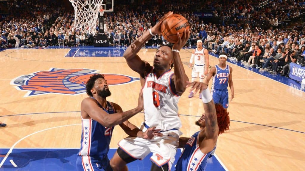 new york lay up 1 1 - Digital Wager Wire Knicks player in white drives to the basket while two defenders reach in during a crowded game on a packed arena court of fans behind him