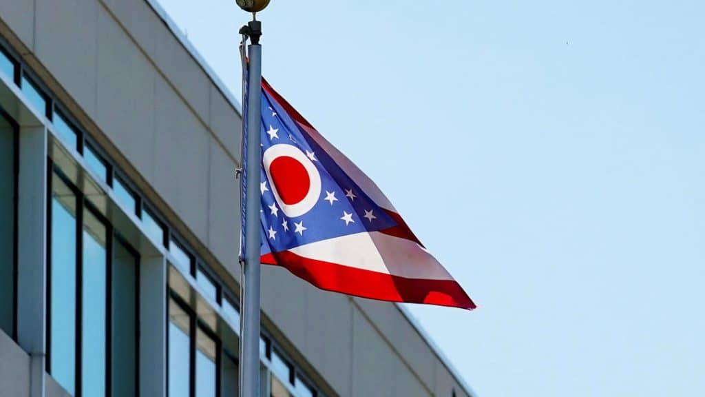 ohio removal of cards 1 - Digital Wager Wire Flag of Ohio waving on a flagpole beside a modern glass building under a clear blue sky.