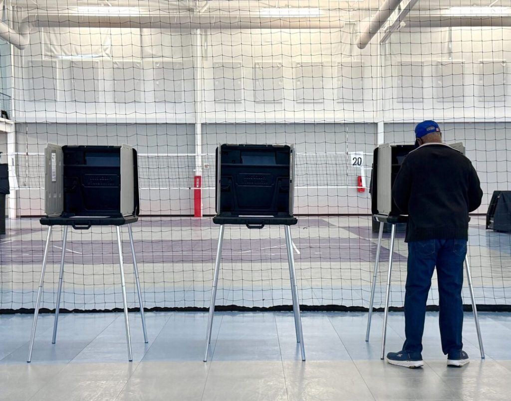 voting 1 - Digital Wager Wire Man wearing a blue cap votes at a privacy booth in a gymnasium, with a mesh divider behind.