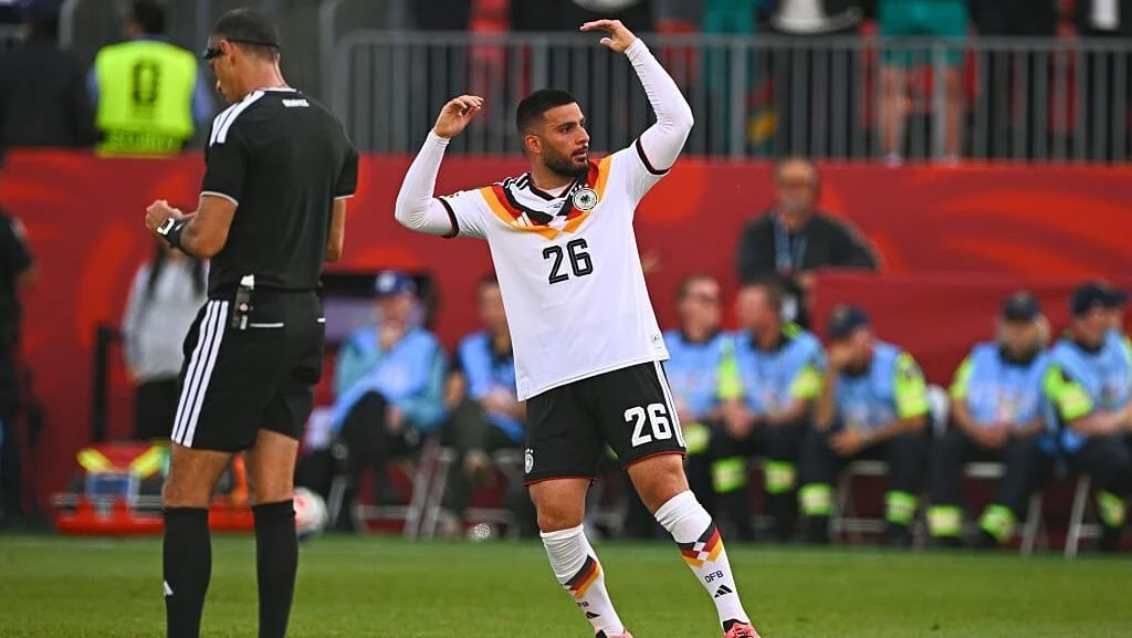 germany raising hands - Digital Wager Wire Soccer player in a white jersey number 26 dancing with arms raised on the field, with staff and spectators on the sideline in the background.
