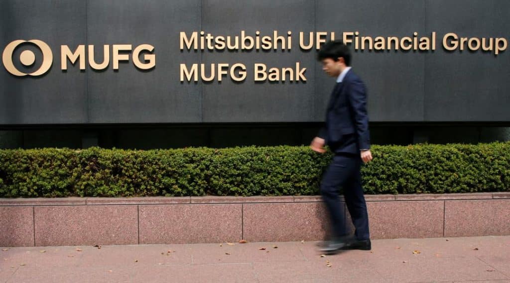 mufg banks - Digital Wager Wire Man in a navy suit walks along a hedge in front of a dark wall with MUFG and Mitsubishi UFJ Financial Group logos visible behind him.