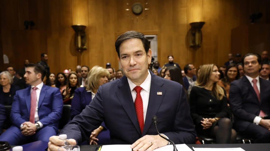 rubio marco - Digital Wager Wire Man in a navy suit and red tie sits at a desk with a microphone in front, in a wood-paneled conference room, audience behind him.