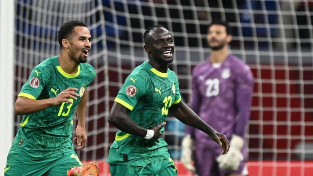 senegal - Digital Wager Wire Two soccer players in green jerseys celebrate a goal near the goal net, with a goalkeeper in a purple kit standing in the background.