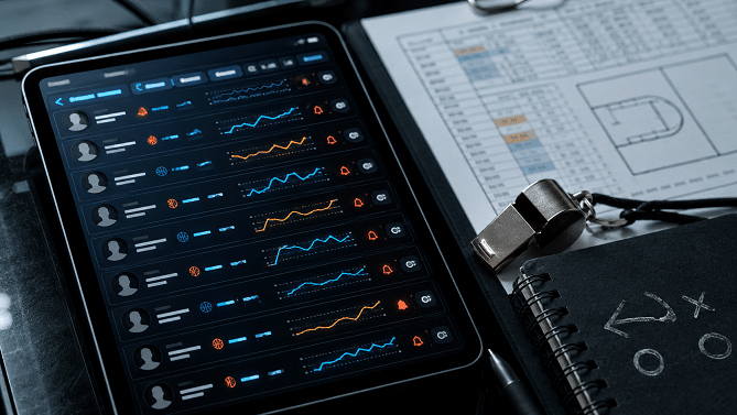 Tablet displaying multiple line charts and user icons over a dark dashboard for data monitoring, with blue and orange lines nearby blueprints and notebook.
