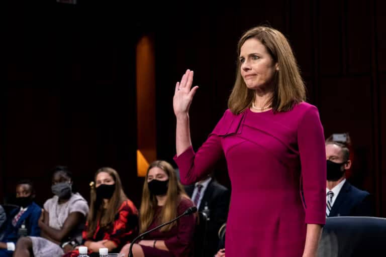 facing senate justice amy - Digital Wager Wire Woman in a magenta dress raises her right hand during a formal oath or procedure in a hearing room, with masked attendees in the background.