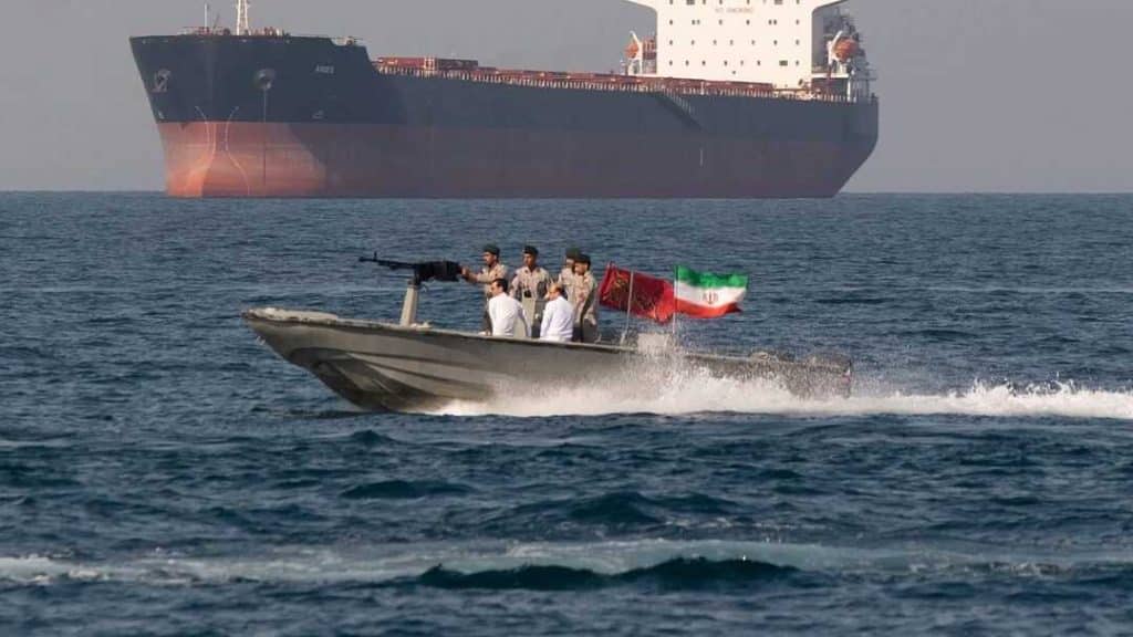 irgc small boat - Digital Wager Wire Motorboat with armed men and an Iranian flag speeds across the sea, a large cargo ship looms in the background.