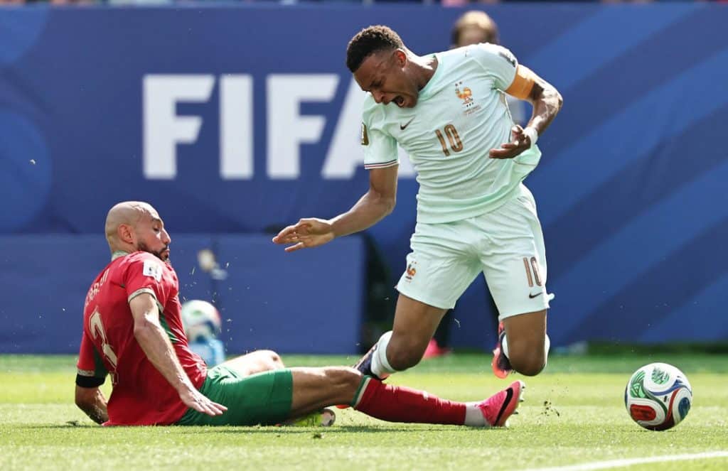 mbappe was trip - Digital Wager Wire Soccer player in a light blue France kit (number 10) leaps as a red-clad opponent slides in on a grassy pitch, ball near them with a FIFA banner in the background.