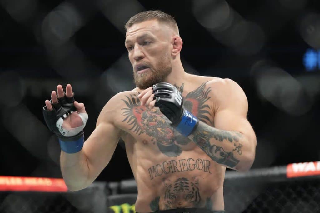 the champ - Digital Wager Wire Tattooed male MMA fighter inside a fenced cage, wearing blue hand wraps and black gloves, mid-fight stance with fists raised.