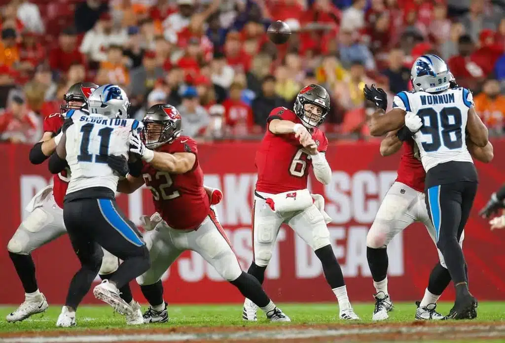 Quarterback in a red jersey throws a pass as defenders in red and white collide near the line of scrimmage, ball suspended in midair.