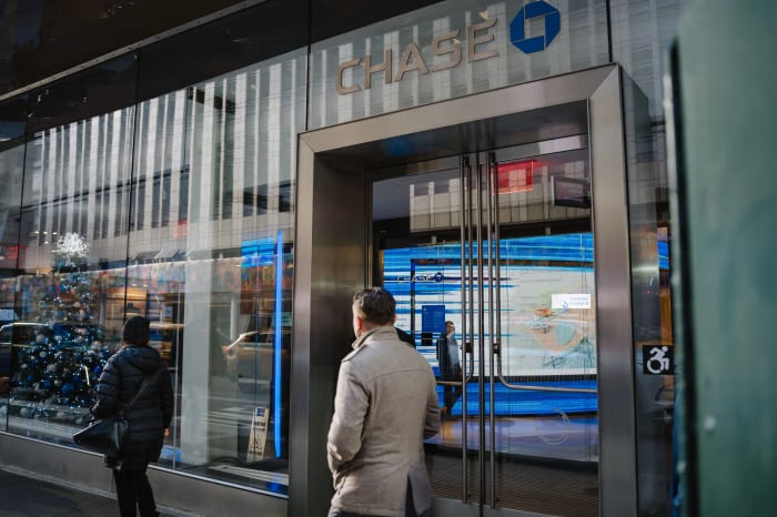 chase bank - Digital Wager Wire Man in a gray coat stands outside the glass entrance of a Chase bank, reflecting city lights and storefronts.