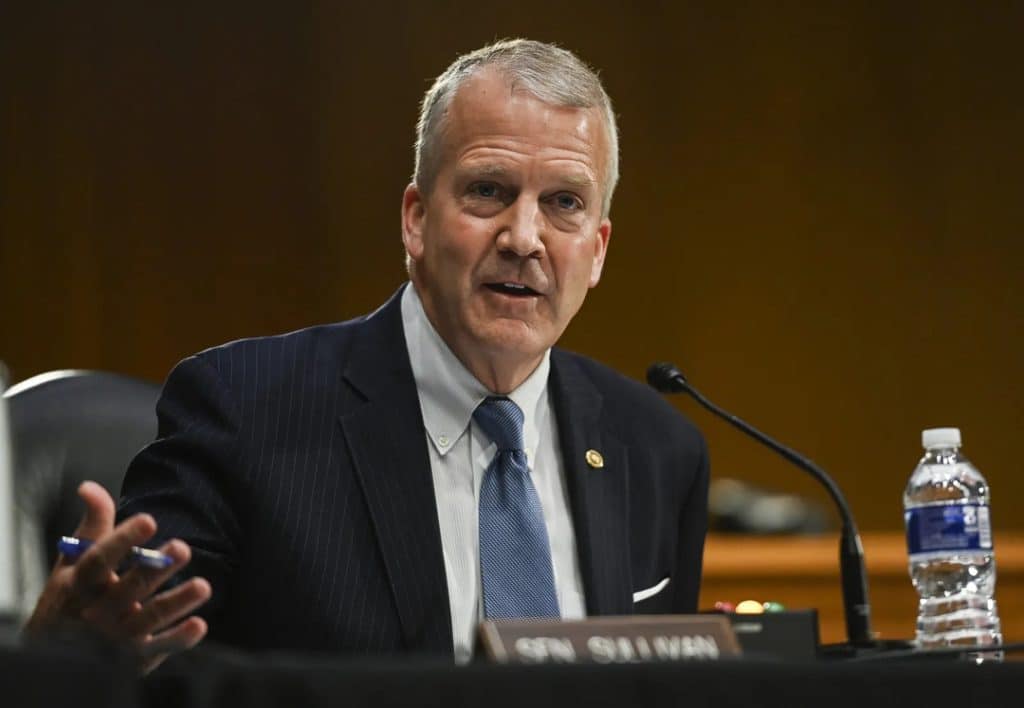 dan sullivan - Digital Wager Wire Middle-aged man in a dark pinstripe suit and blue tie speaks at a formal hearing, with a microphone and water bottle on the table.
