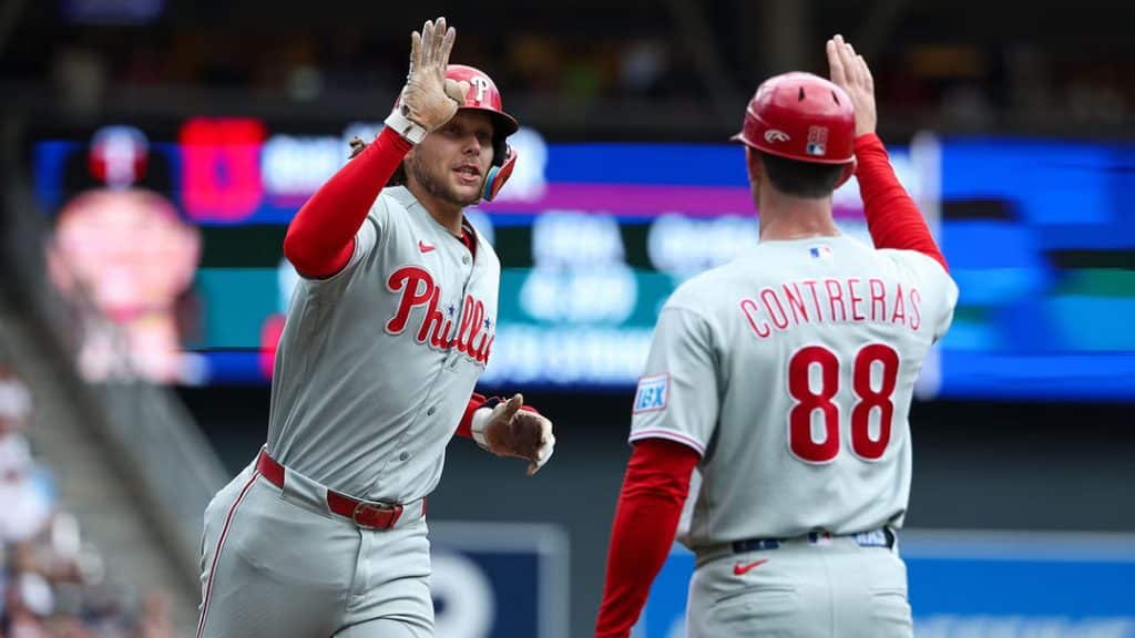 phillies contreras - Digital Wager Wire Two Philadelphia Phillies players high-fiving on the baseball field; one facing the camera in a gray away uniform with red sleeves and helmet on the head.