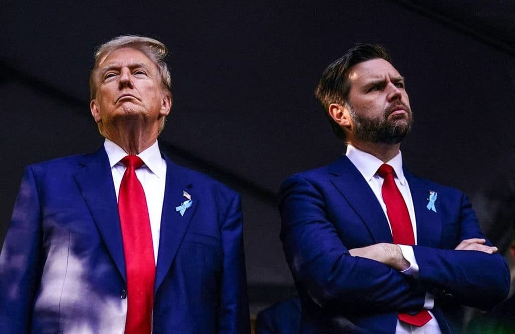 trump and vance on the podium - Digital Wager Wire Two men in blue suits with red ties stand on a dark stage, looking upward with serious expressions; each wears a light blue ribbon on the lapel.
