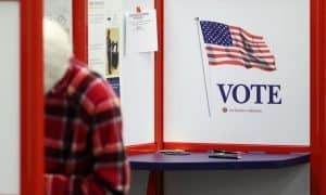early vote - Digital Wager Wire Voting booth with a large 'VOTE' sign and waving American flag; a person in a red plaid shirt stands nearby.